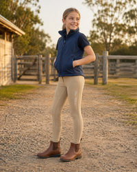 A young girl stands on a gravel path in front of a rustic wooden fence and barn, wearing a navy blue sleeveless zip-up vest, beige riding tights, and brown Chelsea boots.