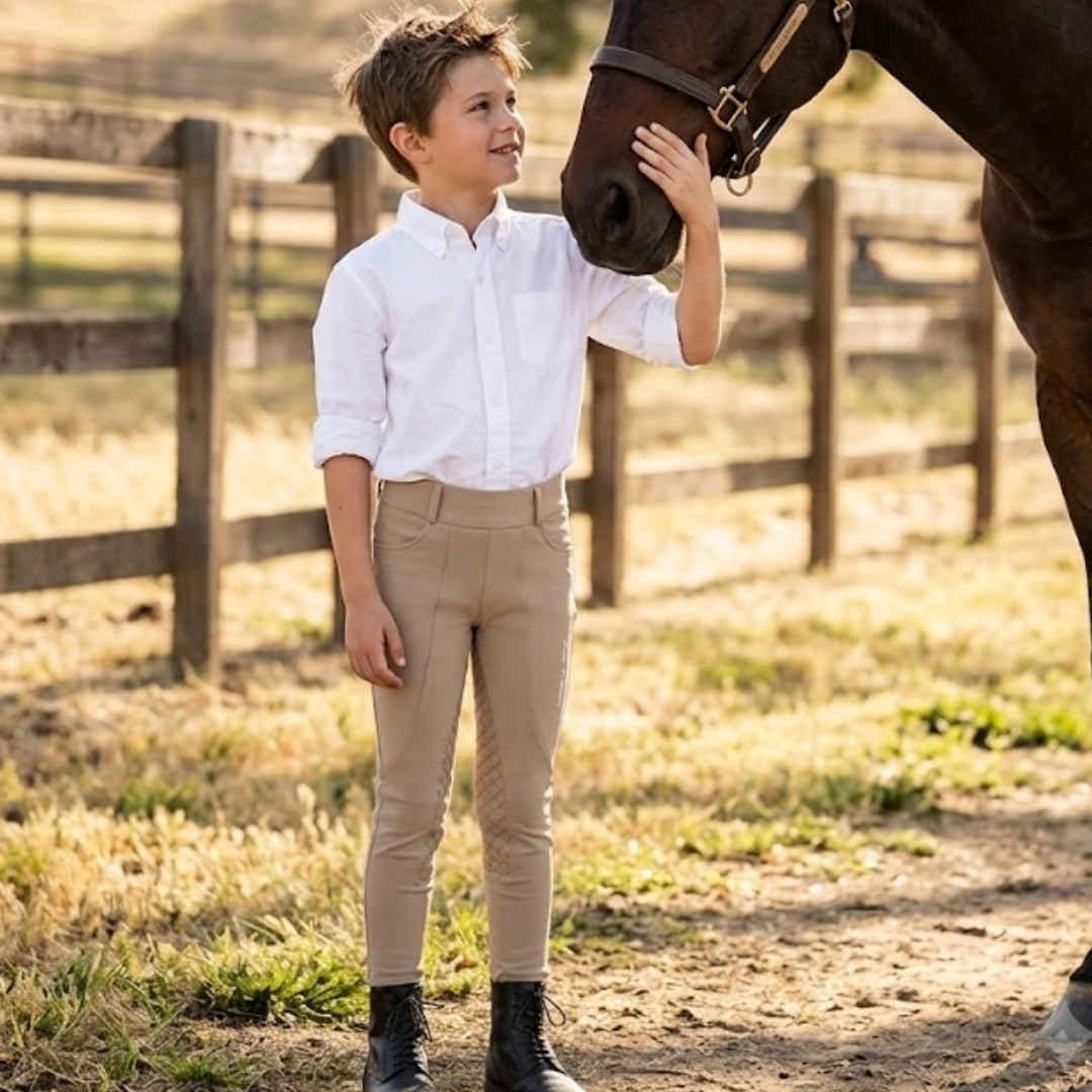 A young boy wearing a white long-sleeve button-down shirt and beige riding breeches stands in a sunlit paddock, gently touching a brown horse's face. The background features a rustic wooden fence and dry grass.