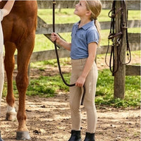 A young girl in a blue polo shirt and beige riding breeches stands in a horse paddock holding a horse lead rope next to a brown horse.